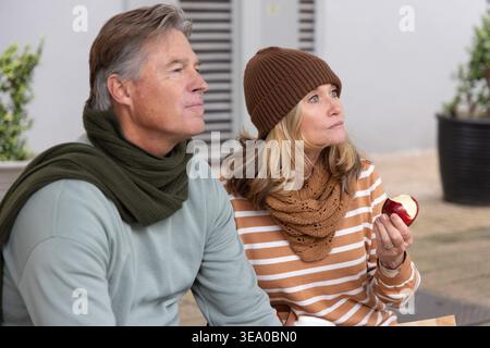 Senior couple sitting on bench in courtyard holding bitten red apple, wearing knit beanie, scarves. Duo, outdoor, plaza, urban, contemplative, greenery, seating Stock Photo