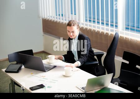Professional businesswoman working on laptop in a bright office space. Focused female employee analyzing reports with a cup of coffee.  Stock Photo