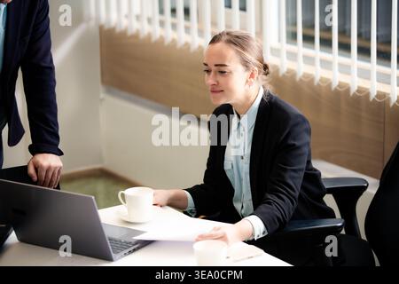 Professional businesswoman working on laptop in a bright office space. Focused female employee analyzing reports with a cup of coffee.  Stock Photo