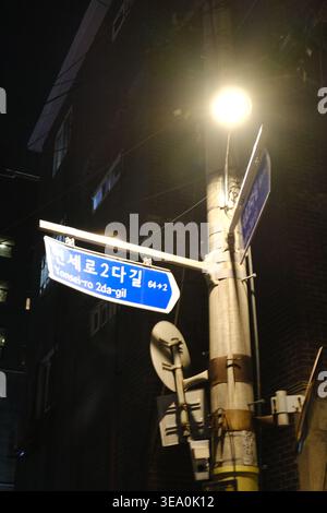 A glowing street lamp illuminating blue Korean road signs (Yonsei-ro 2da-gil) against the dark silhouettes of buildings in a Seoul alleyway at night. Stock Photo