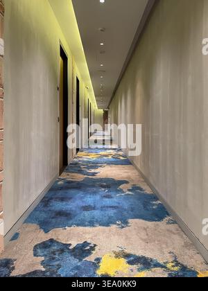 A long, modern hotel corridor in Penang, Malaysia, with a vibrant blue and yellow patterned carpet stretching toward the vanishing point. Warm yellow accent lighting illuminates the grey walls from above, creating a stylish and welcoming atmosphere suitable for hospitality and interior design content. Stock Photo