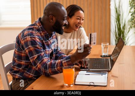 Happy multiracial couple using laptop and payment card to pay financial bills at home. Stock Photo