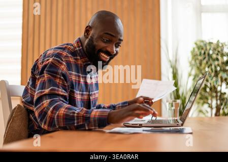 Happy black man is doing home budget accountancy. He is holding financial bills and using laptop. Stock Photo