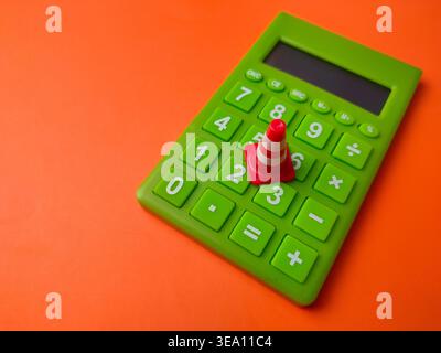 Miniature red and white traffic cone placed on the numeric keypad of a green calculator. Stock Photo