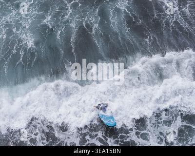 Aerial view of a surfer on a blue board riding through large crashing waves and white sea foam in Yilan County, Taiwan. Stock Photo