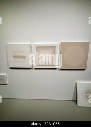 Minimalist art pieces hang on a plain white wall. Three square canvases feature subtle, abstract designs. Soft beige tones contrast with the neutral b Stock Photo