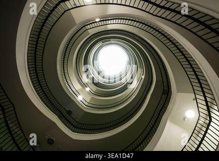 Low angle view of a spiral staircase. An upward perspective of a grand spiral staircase featuring dark railings and a bright skylight. Stock Photo