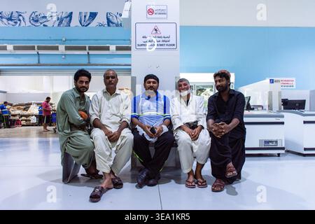 Group portrait of five Asian men in traditional clothing and workwear sitting together on a bench, taking a break inside the Waterfront Market, Dubai. Stock Photo