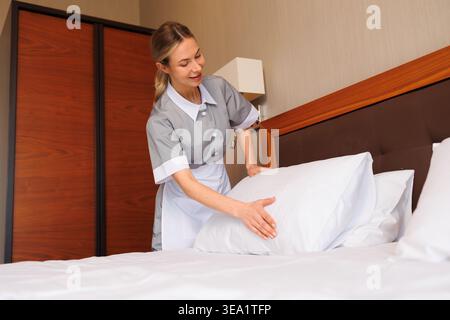 Hotel housekeeper adjusting pillows on comfortable bed in elegant guest room Stock Photo
