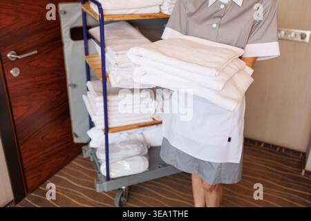 Hotel maid carrying fresh folded towels beside housekeeping cart in corridor room interior Stock Photo