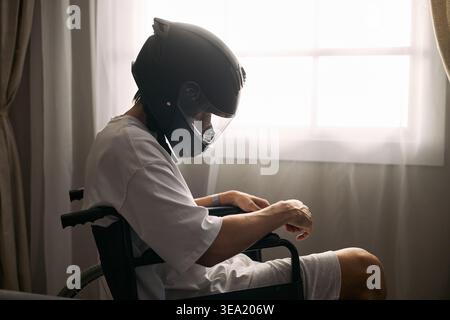 Young adult man with disability sitting in wheelchair wearing motorcycle helmet, suggesting recovery or risk awareness. Useful for insurance, rehabilitation, road traffic safety campaign marketing Stock Photo