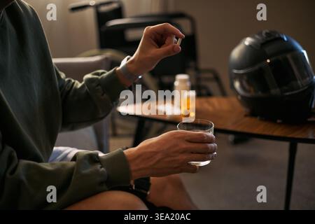 Young adult man with disability holding pill and water glass near wheelchair, taking medication at home. Useful for healthcare, recovery, chronic condition, insurance, caregiving campaigns Stock Photo