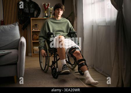 Portrait of Caucasian teenager sitting in wheelchair with leg brace during home recovery after injury, representing rehabilitation and mobility support. Useful for healthcare campaigns Stock Photo