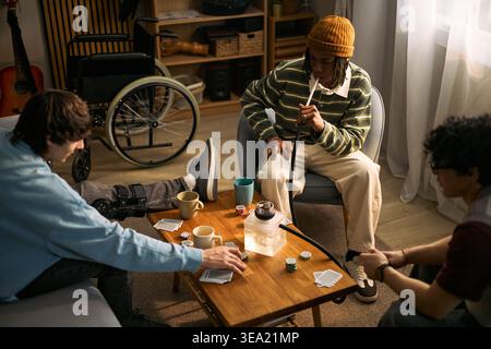 Teenagers with disability socializing and playing cards around coffee table, using wheelchair and leg brace in shared room. Ideal for inclusion, accessibility, youth support campaigns Stock Photo