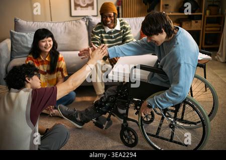 Young adult man with disability in wheelchair high fiving male friend while diverse friends gathering at home, representing inclusion and support. Ideal for accessibility campaigns Stock Photo
