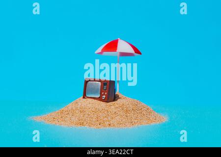 on a small mound of sand, a vintage television rests beside a red and white beach umbrella, isolated against a bright blue background Stock Photo