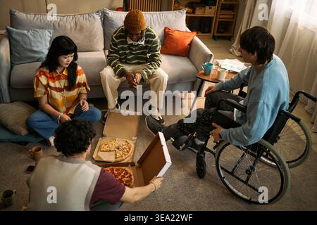 High angle view of diverse young adults sharing pizza during home gathering, including Asian woman and man with disability using wheelchair. Suitable for inclusion and friendship campaigns Stock Photo