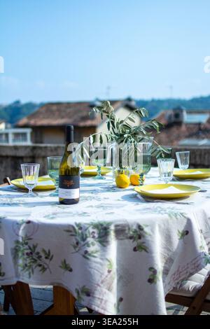 Outdoor dining table set with wine bottle, lemons and olive branches on rooftop terrace with Italian countryside view, Salò, Lake Garda Stock Photo