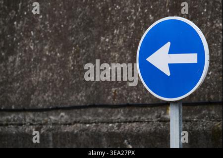 Blue circular mandatory road sign showing a white arrow pointing left, providing clear navigational direction against concrete wall Stock Photo