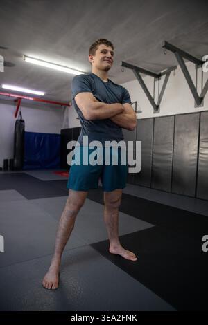 Adult man in sportswear standing barefoot arms crossed on foam mats in gym near heavy bag Stock Photo