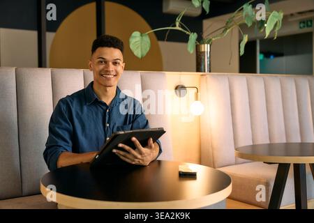 Portrait of cheerful young businessman holding digital tablet while sitting in office lounge at night Stock Photo