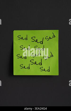 Vertical image of one green sticky note stuck on a dark grey surface that has the word SAD repeated all over it. Stock Photo