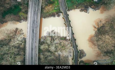 An aerial perspective of a bridge crossing a muddy river, surrounded by trees. A road runs parallel to the bridge. Stock Photo