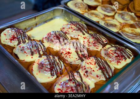 Freshly baked cinnamon rolls with white frosting, chocolate drizzle, and red sprinkles in a metal tray. Sweet pastry in a bakery. Stock Photo