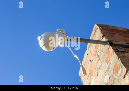 A white plastic CCTV camera mounted on a metal bracket on a brick building under a clear blue sky. Security system, outdoor technology, home protectio Stock Photo