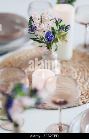 Table decorated with spring flowers in vases and candles for dinner at home. Close up of bouquet of blooms by dinnerware, cutlery Stock Photo