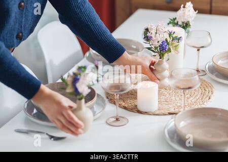 Woman decorating dining table with spring flowers in vases and candles at home. Close up of dinnerware, cutlery served for four.  Stock Photo