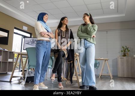 Diverse female colleagues wearing hijab, kurta and sweater collaborating in studio by easel Stock Photo