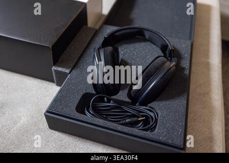 Black over-ear headphones sit in open packaging. Cable lies neatly coiled beside them. Box rests on textured beige surface. Clean, minimalist product Stock Photo