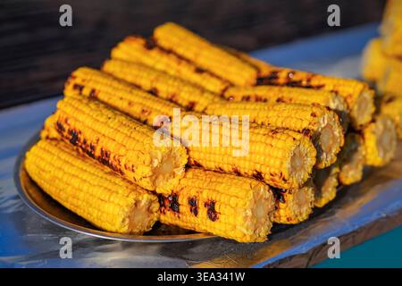 Grilled corn cobs stacked on metal tray with charred marks and bright yellow kernels prepared in outdoor street food market. Street food, grilled corn Stock Photo