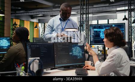 Manager talking with intern in office, using specialized hardware to accelerate complex AI calculations. Team leader teaching trainee how to manage machine learning training datasets, camera A Stock Photo