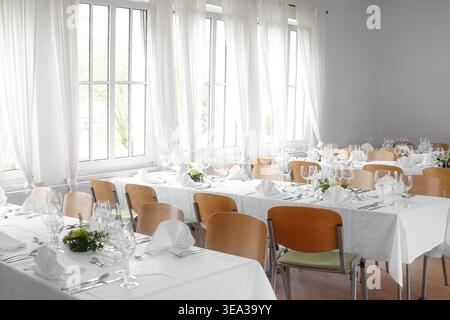 Festive table settings with white tablecloths, folded napkins, glasses and cutlery in a modern meeting room with large windows and bright curtains, se Stock Photo