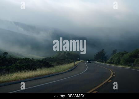 Car driving on winding coastal road through misty hills and low clouds Stock Photo