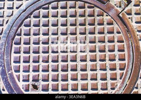 Weathered metal manhole cover shows a grid of raised squares and a small notch along its rim. Iron utility access cover set into the ground Stock Photo
