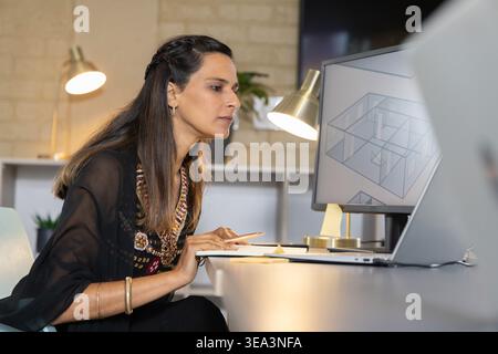 Adult Indian woman wearing embroidered blouse leaning at desk in studio viewing 3D plan on monitor Stock Photo