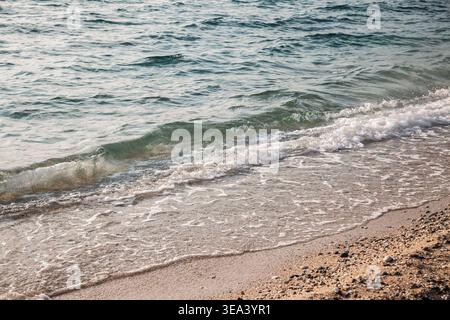 Soft turquoise waves of the Mediterranean Sea roll onto a sunlit shoreline of fine sand and small pebbles. The image captures gentle surf, sea foam an Stock Photo