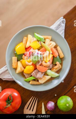 A close-up of stir-fried pork with sweet and sour sauce served in a dish with seasoning and a fork on a wooden table. Top view. Flat lay. Stock Photo