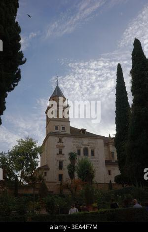 Church of Santa Maria de la Alhambra with ornate bell tower and conical spire rising above garden, framed by cypress trees with tourists below Stock Photo
