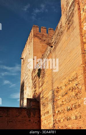 Low-angle view of ancient Alhambra fortress walls and battlemented tower with mixed rubble and brick masonry glowing in warm golden sunlight, Granada Stock Photo