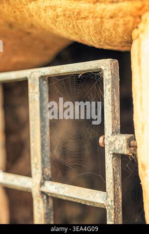 A delicate spider web contrasts with a weathered metal gate set in a rough stone wall, evoking a sense of age and abandonment. Stock Photo