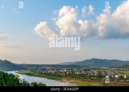 High angle view of a picturesque Chinese rural town along a river valley, surrounded by green mountains under a dramatic summer sky with cumulus cloud Stock Photo