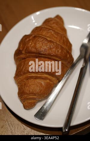 Golden-brown croissant on white plate with knife and fork, placed on wooden surface. Stock Photo