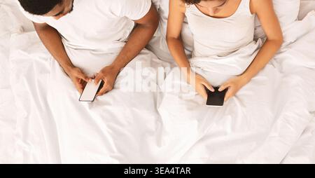 Couple Using Smartphones While Sitting on Bed With White Sheets in the Evening Stock Photo