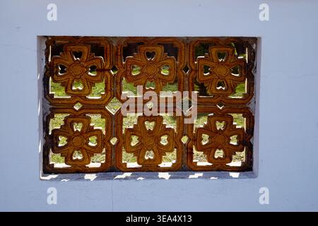 Decorative ventilation block set into a white wall with floral geometric pattern, allowing light and view through to greenery outside. Stock Photo