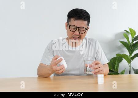 Middle-aged Asian man winces while checking a medicine bottle and holding a glass of water, portraying illness, headache, medication use, and home rec Stock Photo