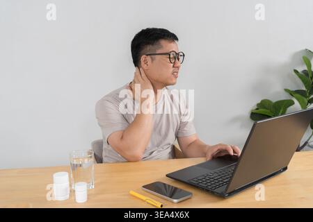 Asian man rubbing his neck while working on a laptop at a desk, with water and medicine nearby, conveying office strain, poor ergonomics, stiffness, a Stock Photo